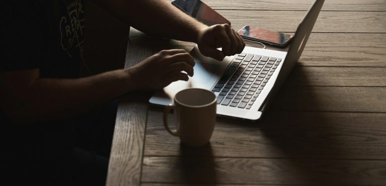 Eine Hand tippt auf einer Laptop-Tastatur neben einer Kaffeetasse und einem Smartphone.